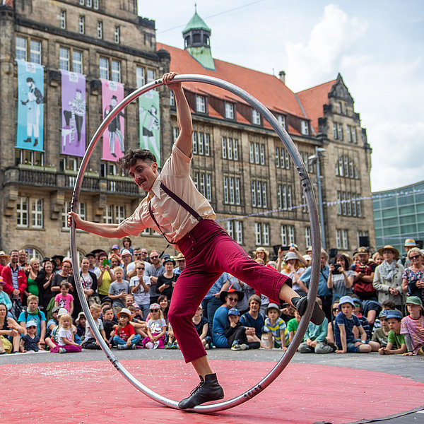 Performer in red pants and suspenders balances inside a large hoop on a red mat, with a crowd watching in front of a historic building.
